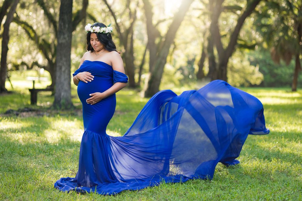 Pregnant woman in flowing royal blue off-shoulder gown with fabric billowing in sunlit green park setting.