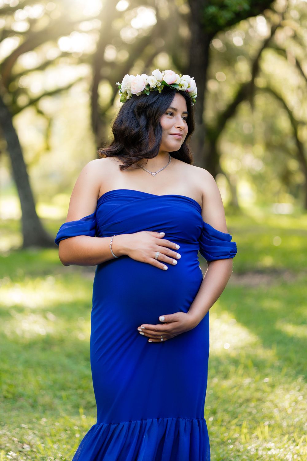 Pregnant woman in royal blue off-shoulder gown wearing floral crown poses outdoors in sunlit park.