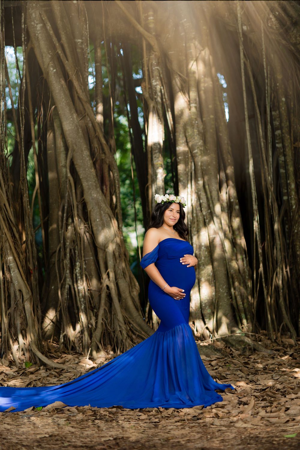 Pregnant woman in royal blue off-shoulder mermaid gown with floral crown poses among banyan trees.