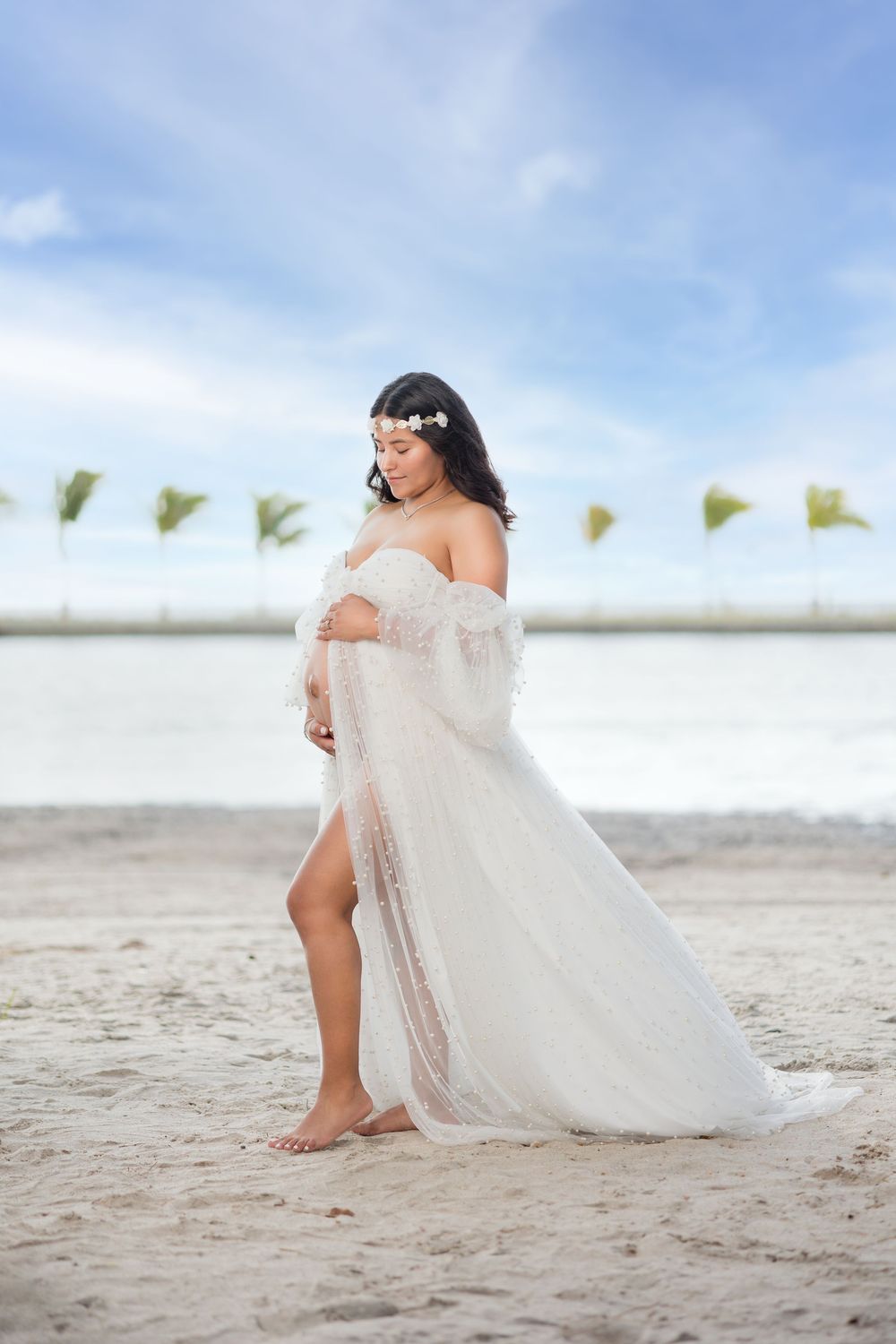 Pregnant woman in flowing white lace gown with floral headpiece stands barefoot on sandy beach.