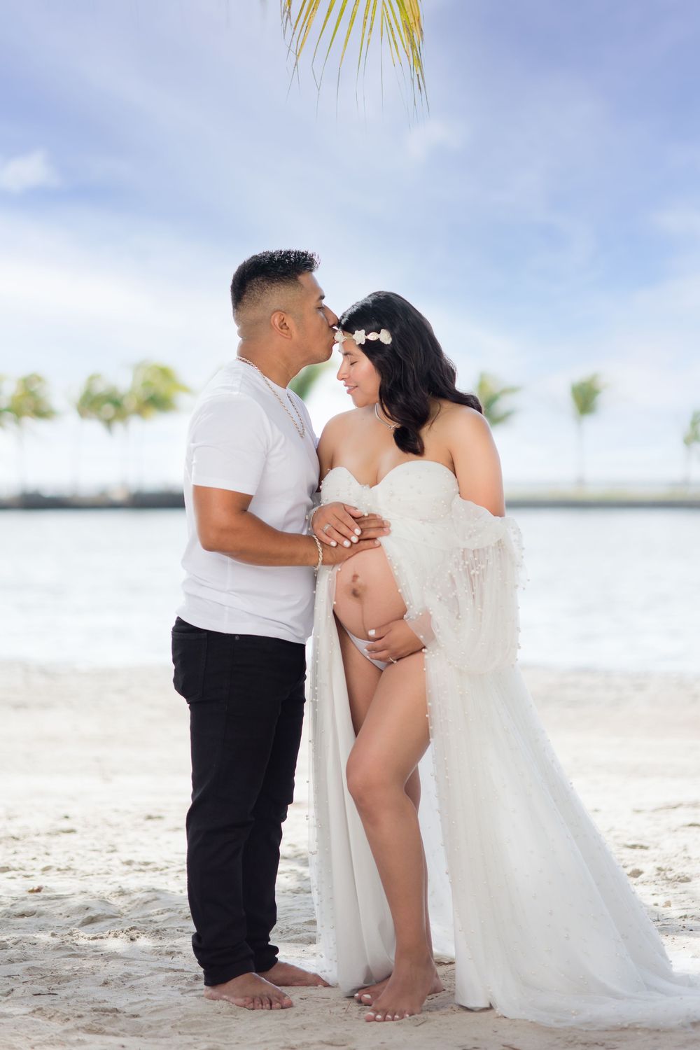 Couple sharing a tender moment on the beach, pregnant woman in white lace gown with floral crown.