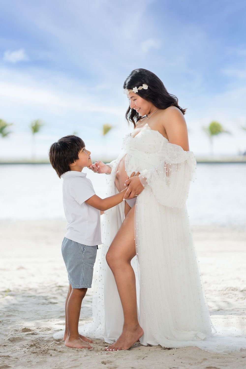 Young boy gently touches pregnant mothers belly as she smiles in white lace gown on beach.