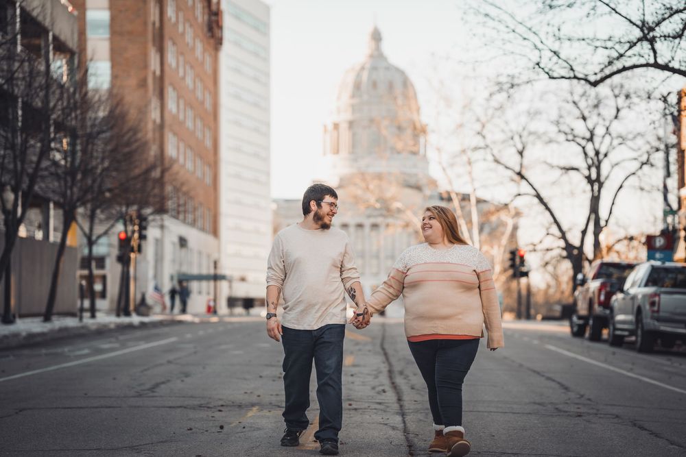 Couple walking together on urban sidewalk with city buildings in background.