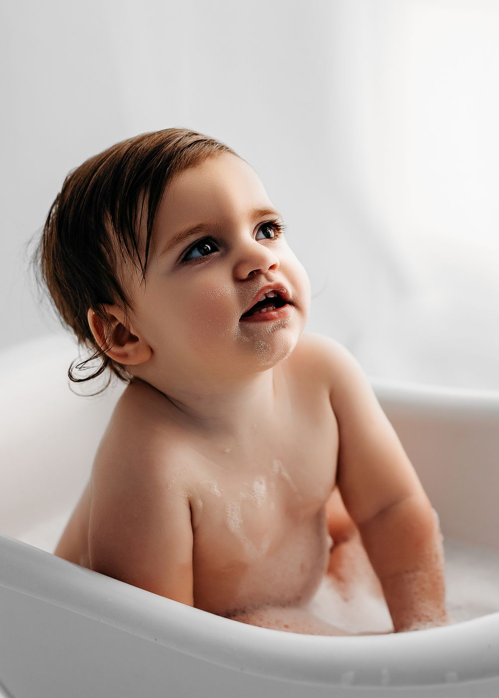 Sweet baby looks up while taking a bath in white bathtub with soft natural lighting.
-Nova photographer- DMV DC during a session in Fairfax,