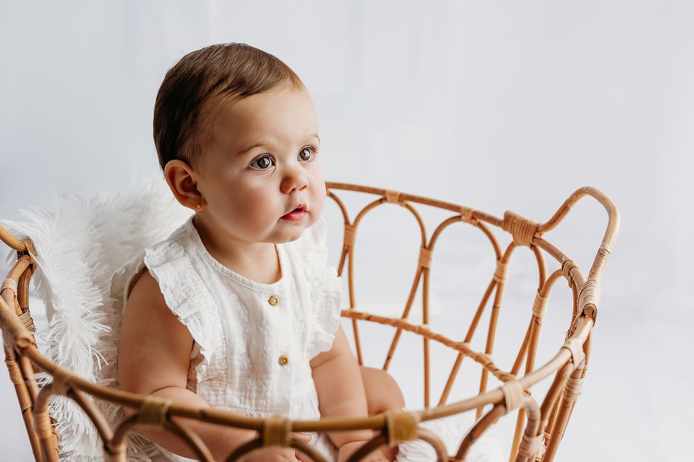 Baby in white dress sits in vintage rattan chair during professional photoshoot.