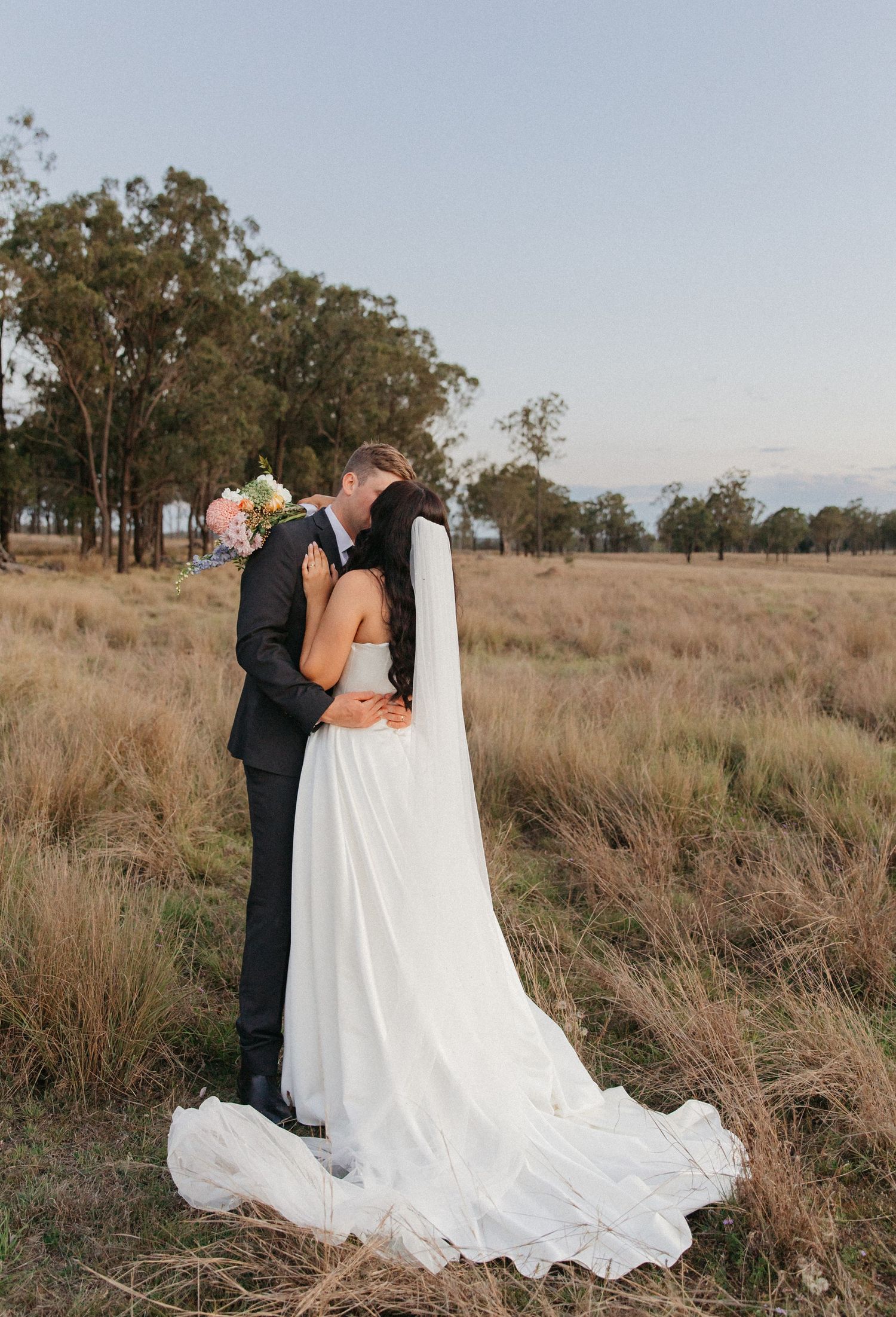 Bride and groom kissing in golden field at dusk, bride wearing long white gown with flowing veil.