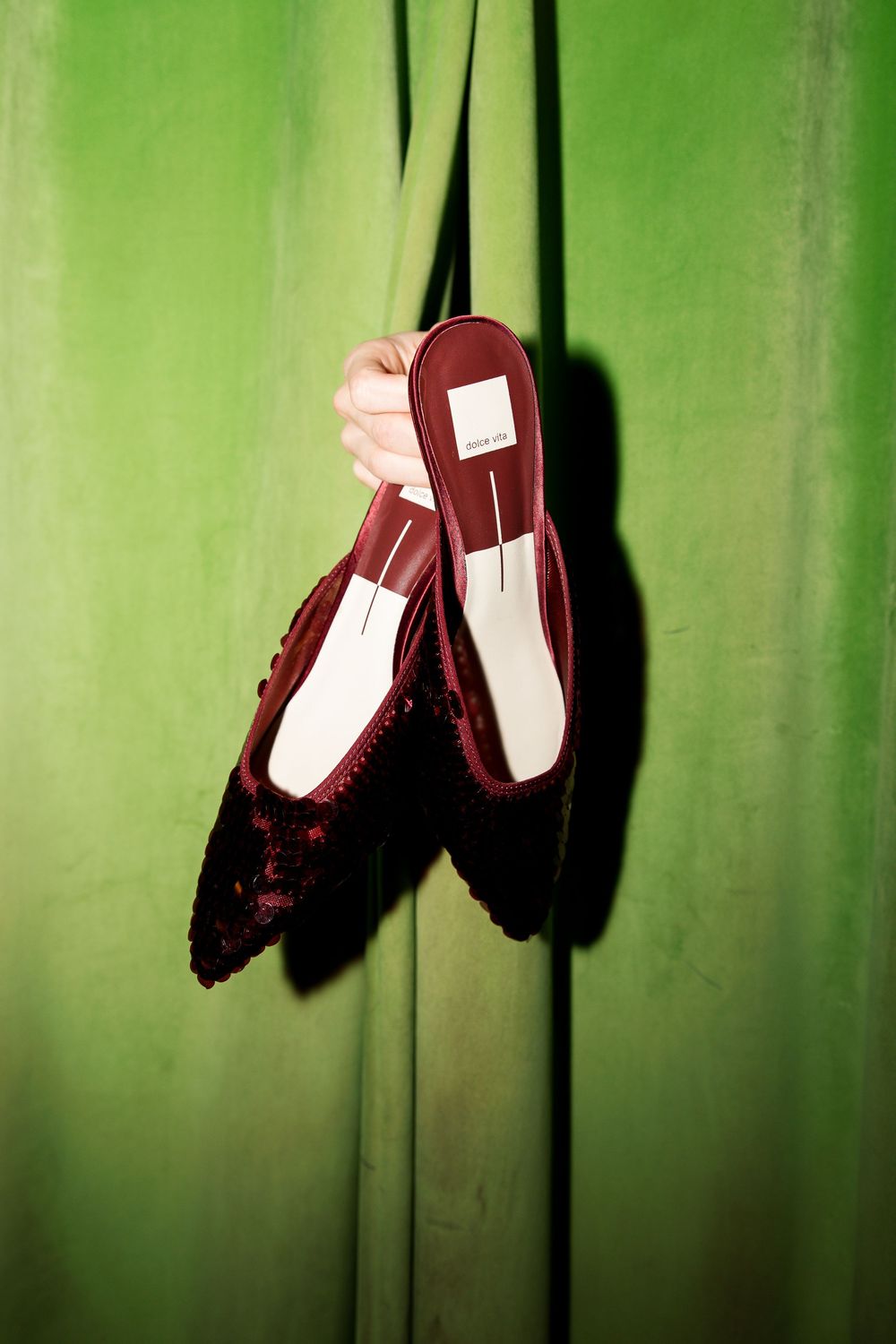 A hand holds burgundy velvet mules with white insoles through a green curtain gap.