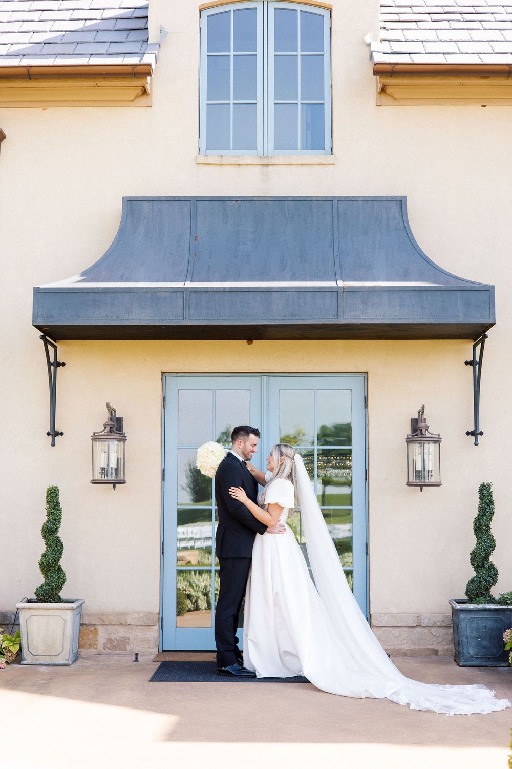 A couple stands beneath a metal awning with lanterns at a French country-style entrance.
