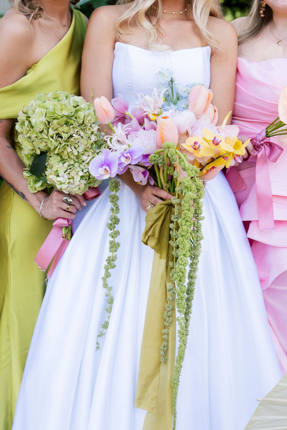 Bride in white strapless gown holds colorful cascading bouquet flanked by bridesmaids in green and pink dresses.