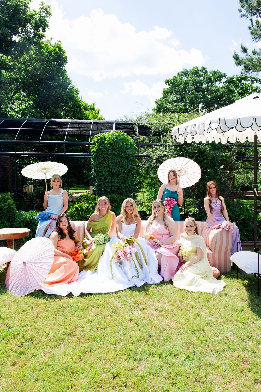 Bride and bridesmaids in colorful dresses posing with parasols in a garden setting on a sunny wedding day.