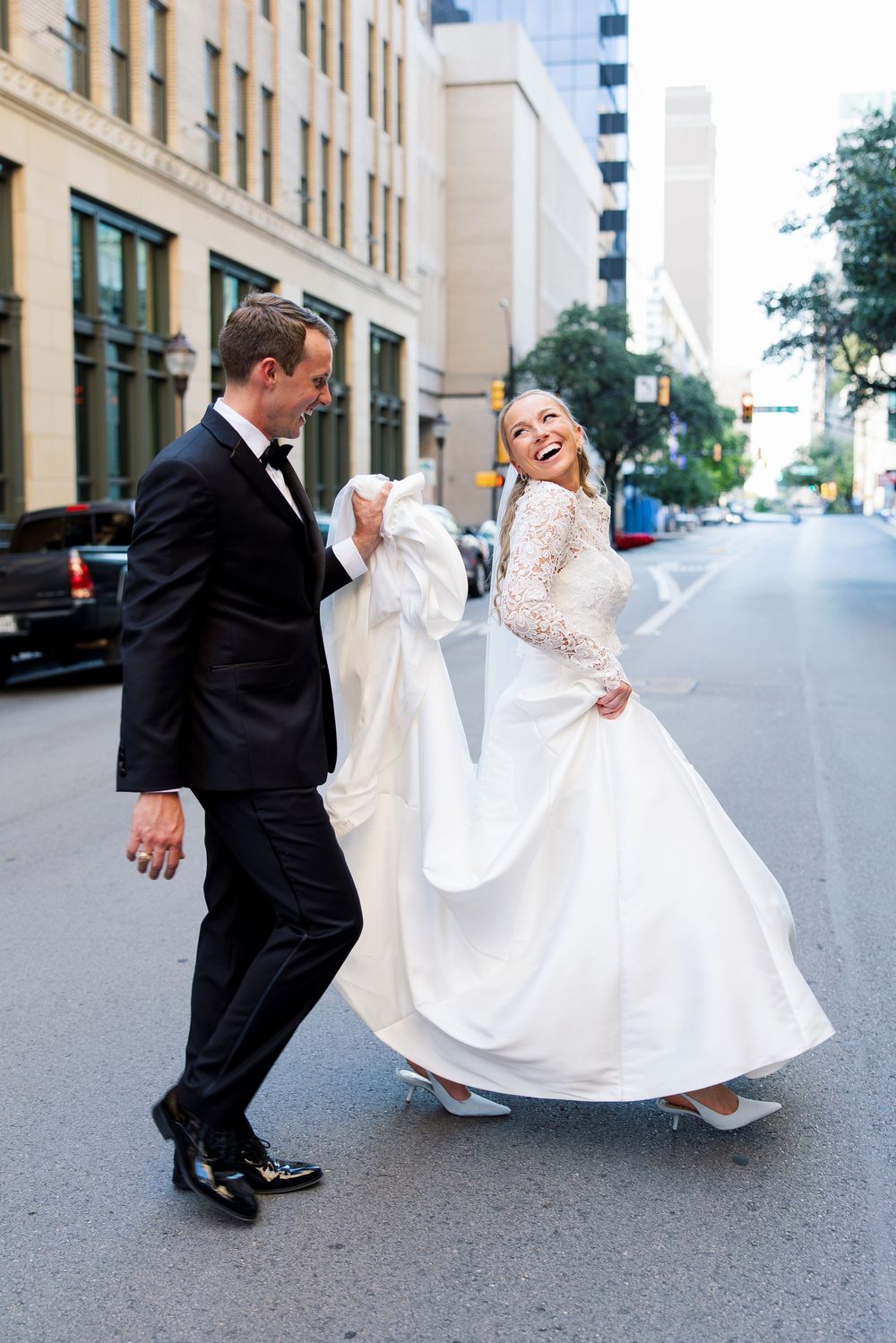 A joyful bride in a white lace gown and groom in black tuxedo walk together on a city street.