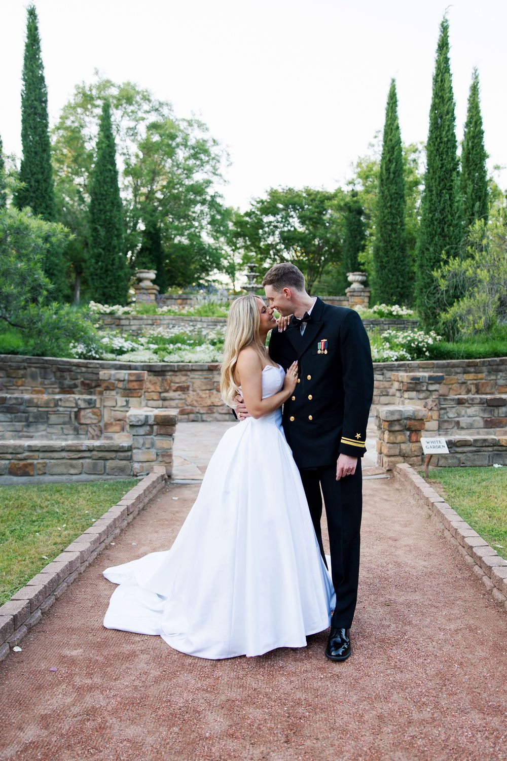 Military groom in dress uniform embraces bride in white ballgown in elegant garden wedding portrait.