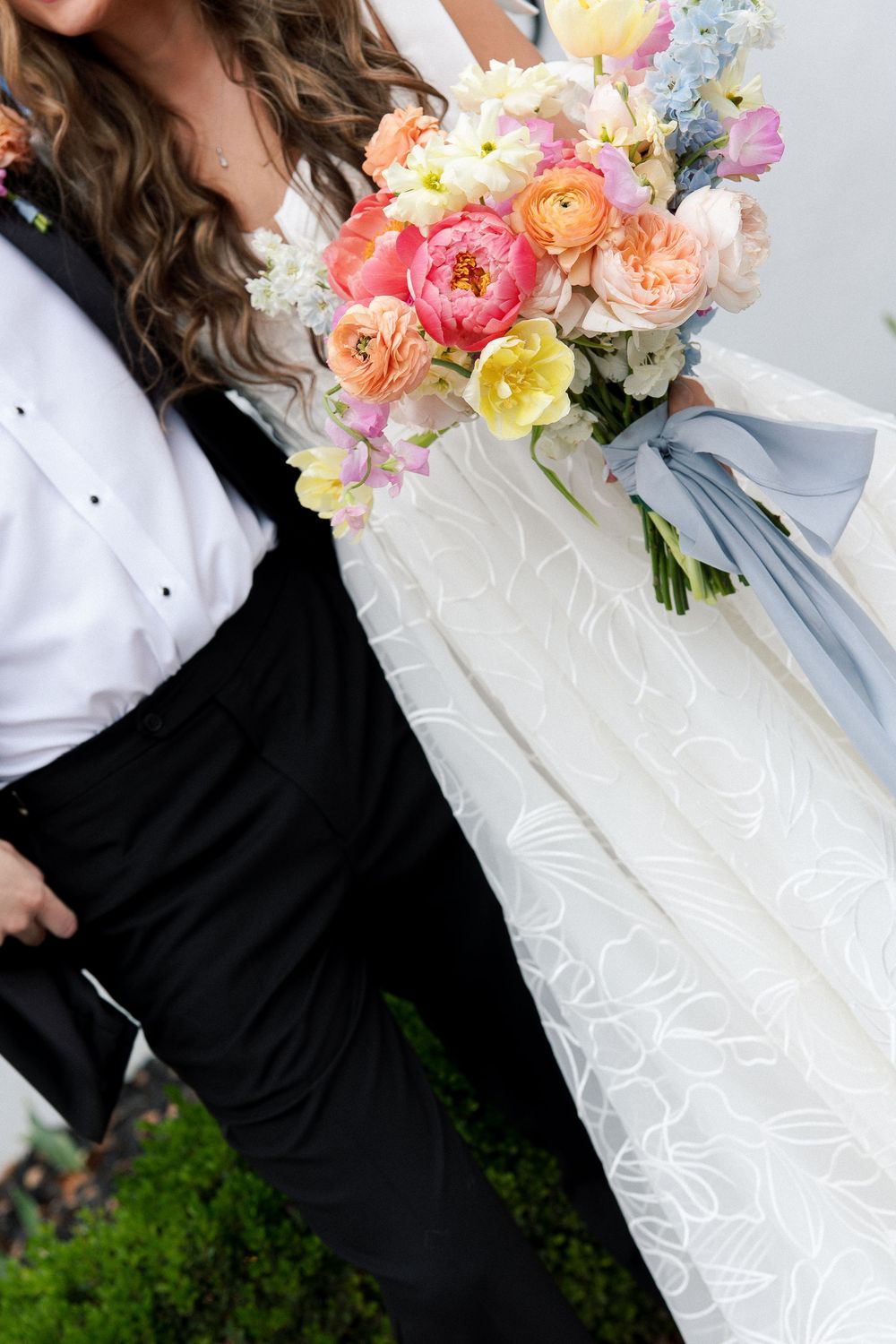 Wedding couple holding a vibrant spring bouquet with coral, yellow and pink flowers against formal attire.