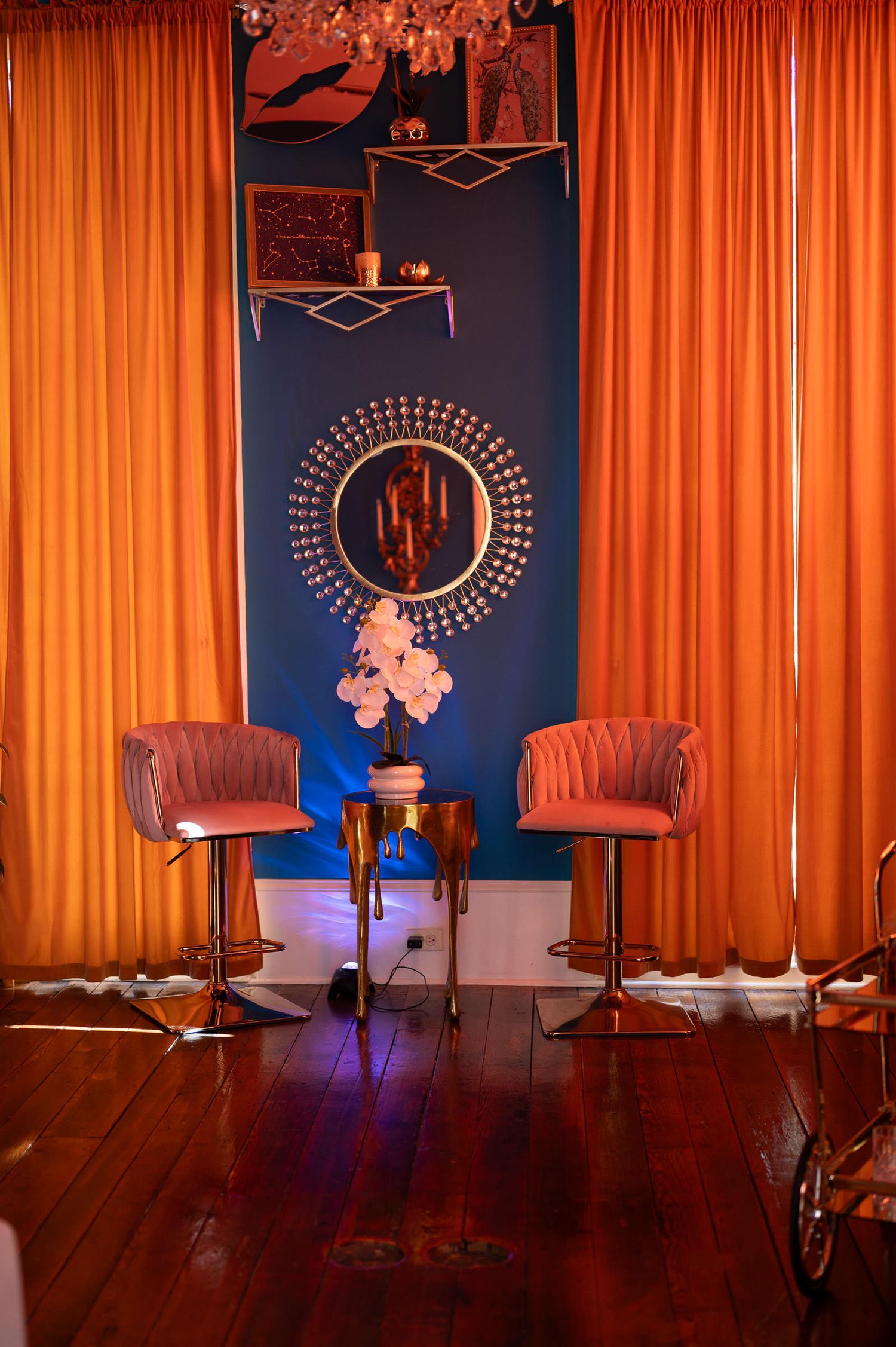 Stylish lounge area with orange curtains, blue accent wall, decorative circular mirror, and two velvet bar chairs.
