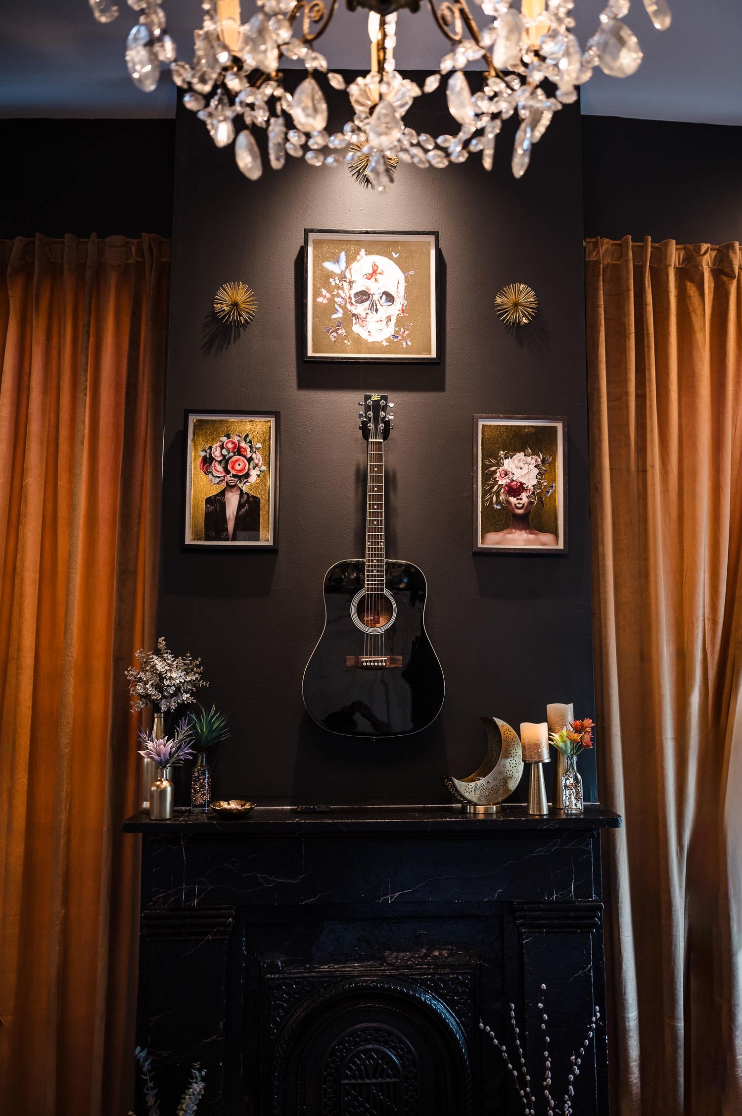 A dark room with orange curtains features crystal chandeliers, guitars on display, and framed artwork on the walls.