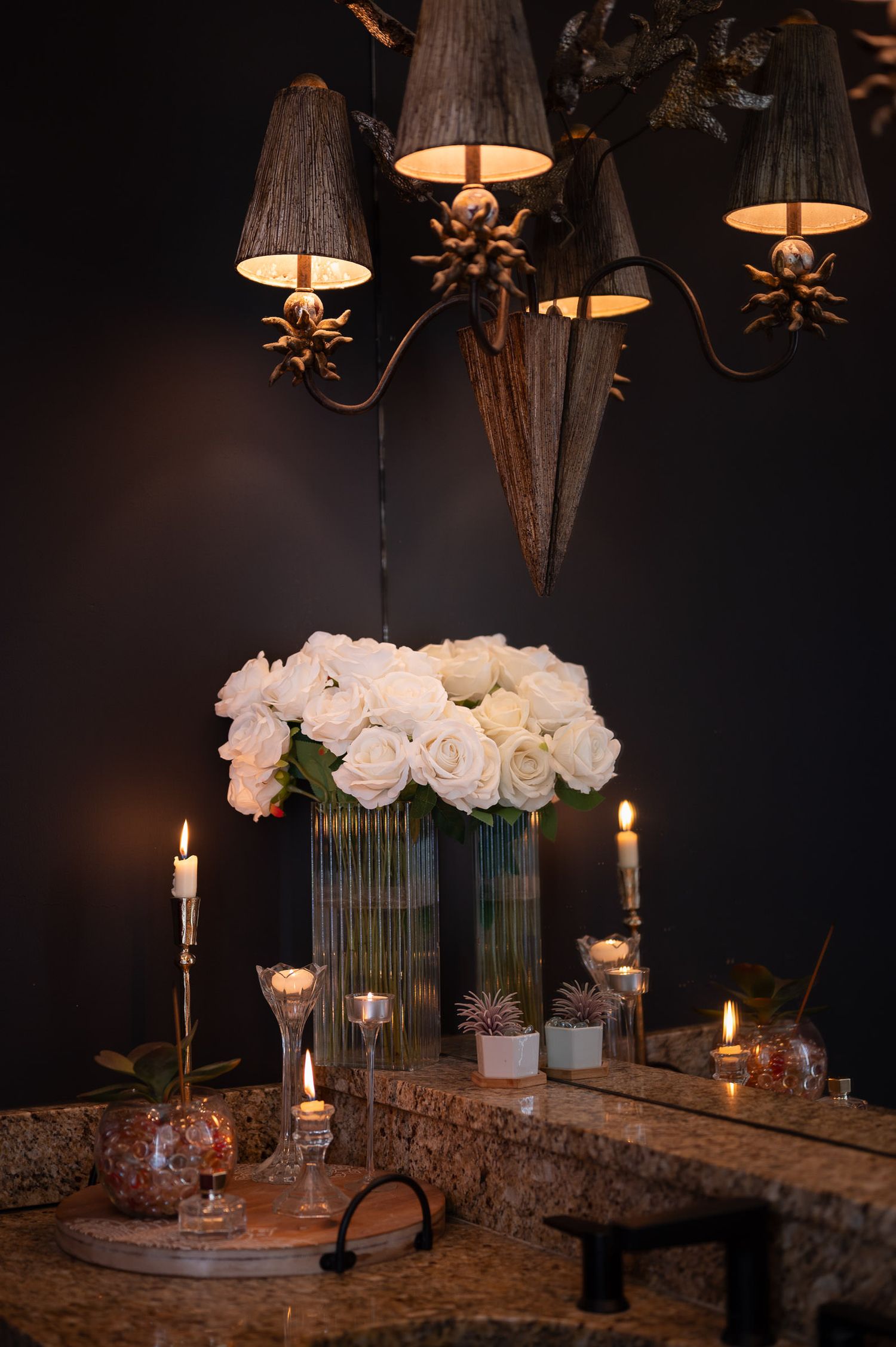 White roses in glass vases illuminate an elegant table setting with glowing candles and dark chandeliers above.