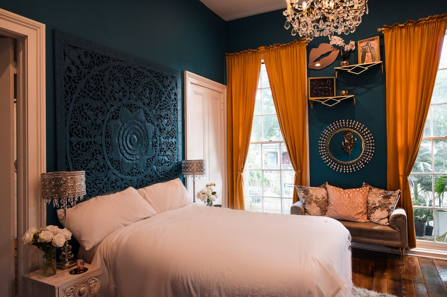 A luxurious bedroom with teal walls, orange curtains, white bedding, and an ornate chandelier creates a dramatic atmosphere.