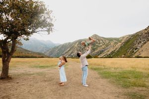 Family enjoying outdoor mountain landscape, father lifting child while mother watches near a tree.