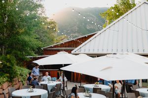 Outdoor patio dining area with white umbrellas, string lights, and mountain views at a rustic venue.