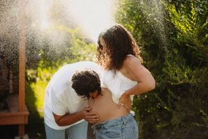 A man kisses a pregnant woman's belly outdoors in golden sunlight, surrounded by lush green trees.