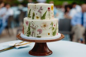 Two-tier white wedding cake decorated with pressed wildflowers and botanical prints on a wooden cake stand.
