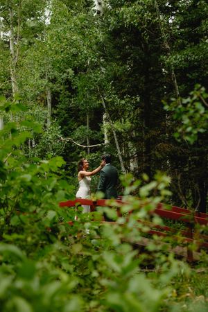 Bride and groom dancing on a red bridge surrounded by lush green forest trees during an outdoor wedding ceremony.