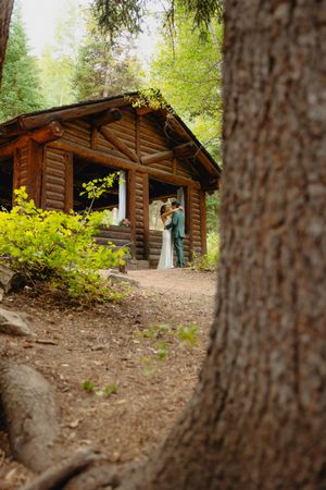 Bride and groom share a romantic moment outside a rustic log cabin surrounded by lush forest trees.