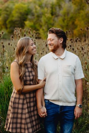 A smiling couple holds hands in a field, woman in plaid dress, man in cream shirt and jeans.