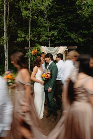 Bride and groom share an intimate moment at outdoor forest wedding ceremony, surrounded by blurred bridesmaids in taupe dresses.