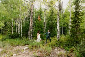 A bride and groom hold hands while walking through a lush aspen forest during their outdoor wedding celebration.