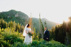 Bride and groom stand among lush green mountain meadow at golden hour, surrounded by pine trees and rocky peaks.