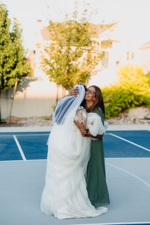 Bride in white gown and veil embracing a woman in green dress on a blue outdoor court on a sunny day.
