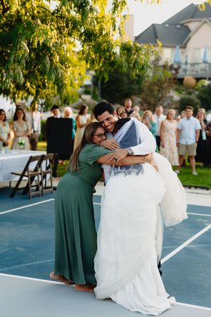 Bride and groom share a joyful group hug with a woman in green dress at outdoor wedding reception.
