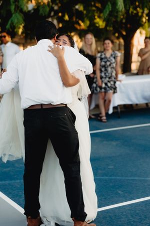 Bride and groom share first dance outdoors on blue court with wedding guests watching in background.