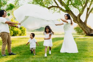 Family playing outdoors on green grass, waving a white blanket over two young children near a large tree.
