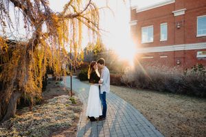 Couple sharing a romantic kiss at golden hour beside a weeping willow tree on a brick pathway.