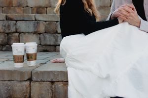 Bride and groom sitting together on stone steps with two coffee cups beside them on their wedding day.