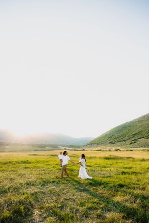 Family walking in a sunlit green meadow with mountains, man holding child, woman in white dress, golden hour light.