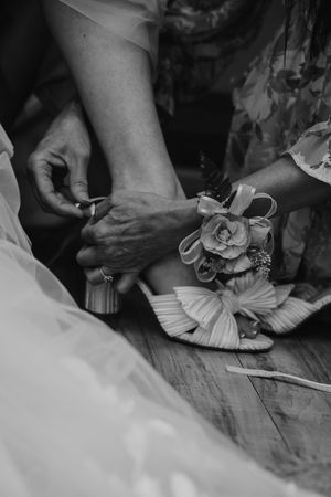 Black and white photo of hands fastening a floral embellished bridal heel on a wooden floor.