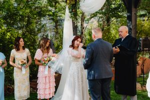 Emotional bride wiping tears during outdoor wedding ceremony with bridesmaids and officiant in lush garden setting.