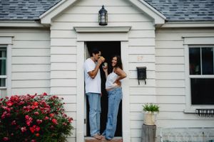 Couple standing in doorway of white home holding a small dog with red roses blooming nearby.