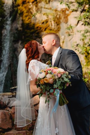 Bride and groom sharing a romantic kiss near a waterfall, bride holding a colorful bouquet in a lace wedding dress.