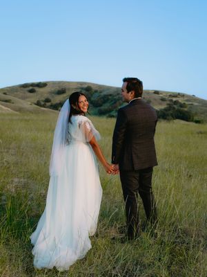 A bride in a white gown and veil holds hands with a groom in a dark suit in a green hillside field.