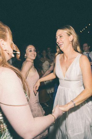 Bride in white gown laughing and dancing with guests in pink dresses at an outdoor wedding reception.