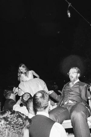 Black and white photo of bride and groom being lifted by guests during lively wedding reception celebration.
