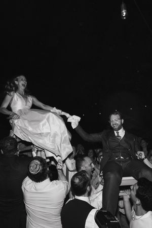 Black and white photo of bride and groom being lifted on chairs during a joyful Jewish wedding hora dance celebration.