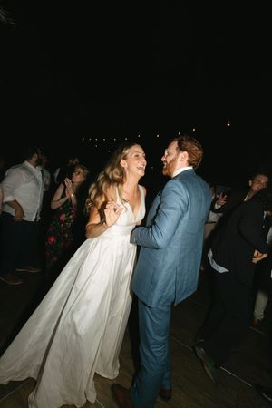 A bride in a white gown and groom in a blue suit share a joyful first dance at their wedding reception.