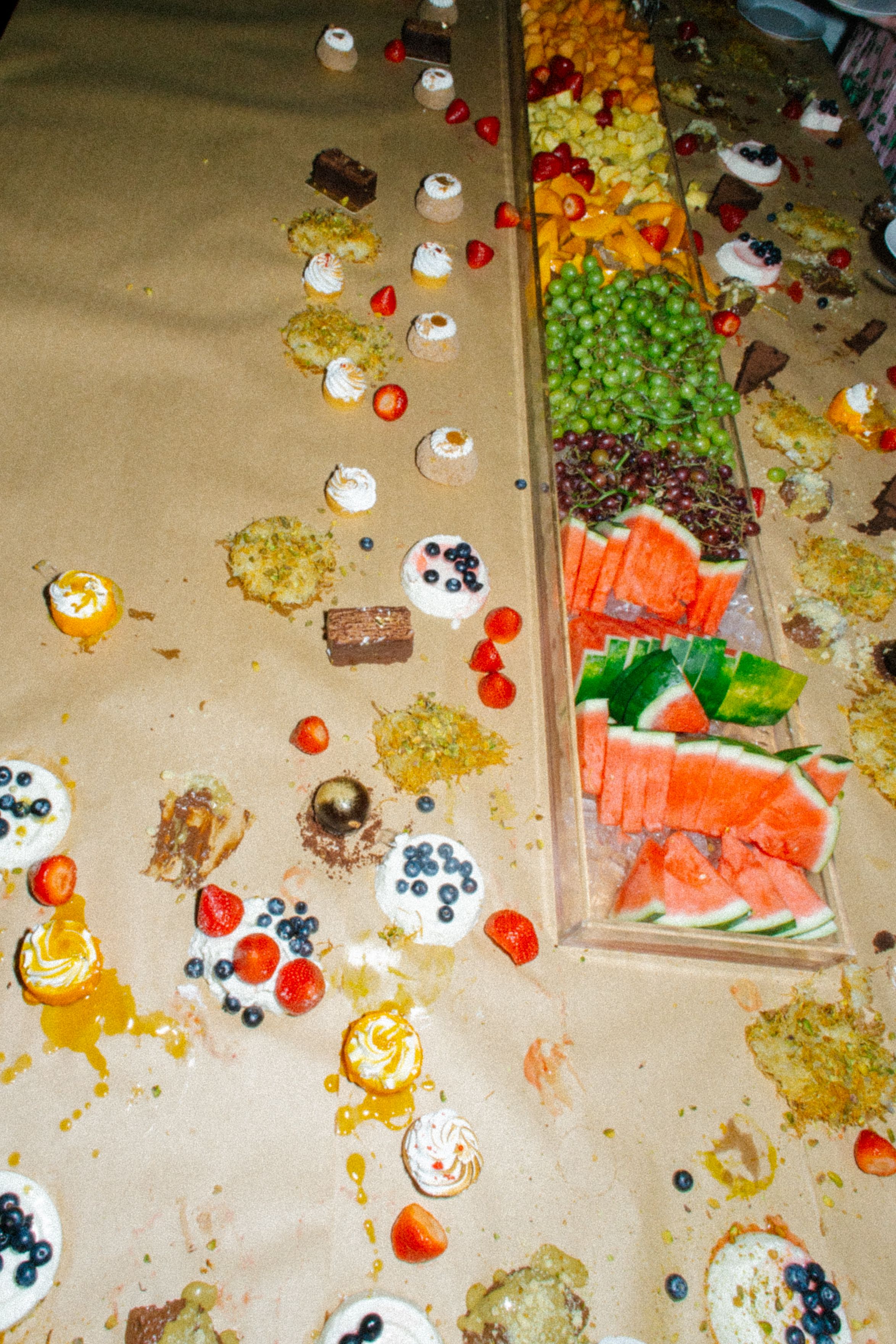 Messy dessert table with scattered fruits, berries, pastries, and watermelon slices after a catered event.