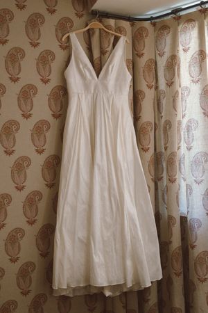 Elegant ivory sleeveless V-neck wedding gown hanging on a wooden hanger against paisley curtains.