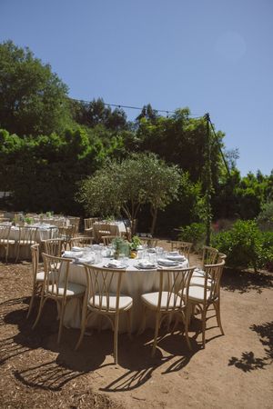 Elegant outdoor wedding reception with round tables, white linens, and wooden chairs set among lush green trees.